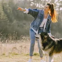 Kot ceketli bir kadının elini yana doğru uzatıp işaret ettiği, yanında duran tüylü siyah-beyaz bir köpeğin fotoğrafı